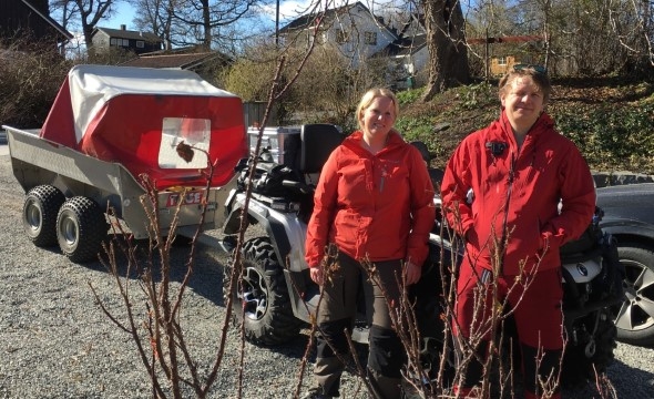 To personer som står i turklær foran en firehjuling som har tilhenger.