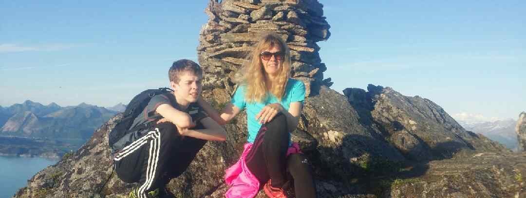 En mor og sønn sitter på toppen av et fjell, ved en varde, og soler seg i fint vær