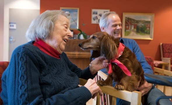 En eldre dame har en svart hund på fanget sitt som hun koser med. Ved siden av henne sitter en annen dame som eier hunden og er frivillig i Røde Kors.