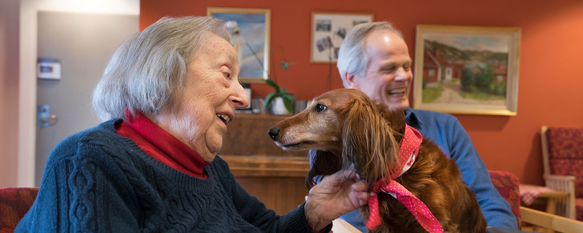 En eldre dame smiler bredt. Hun har en liten brun hund på fanget som hun klapper. I bakgrunnen ser vi en eldre mann som også smiler. 