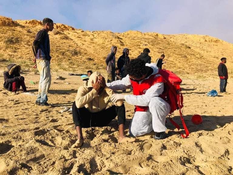 En mann sitter på en strand med hendene over hodet og blir trøstet av en frivillig fra Libya Røde Halvmåne