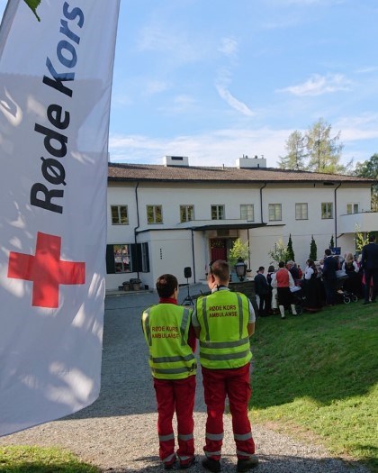 Et Røde Kors banner på siden med to personer fra Røde Kors hjelpekorps. I bakgrunnen ser man en samling av folk foran et hus.