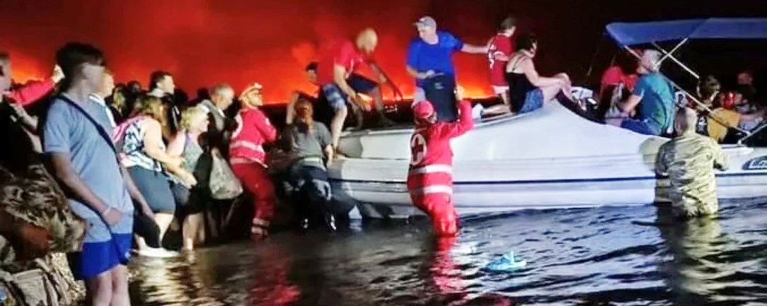 Turister står i vannet og venter på å bli evakuert. En båt med Røde Kors-frivillige henter turistene.