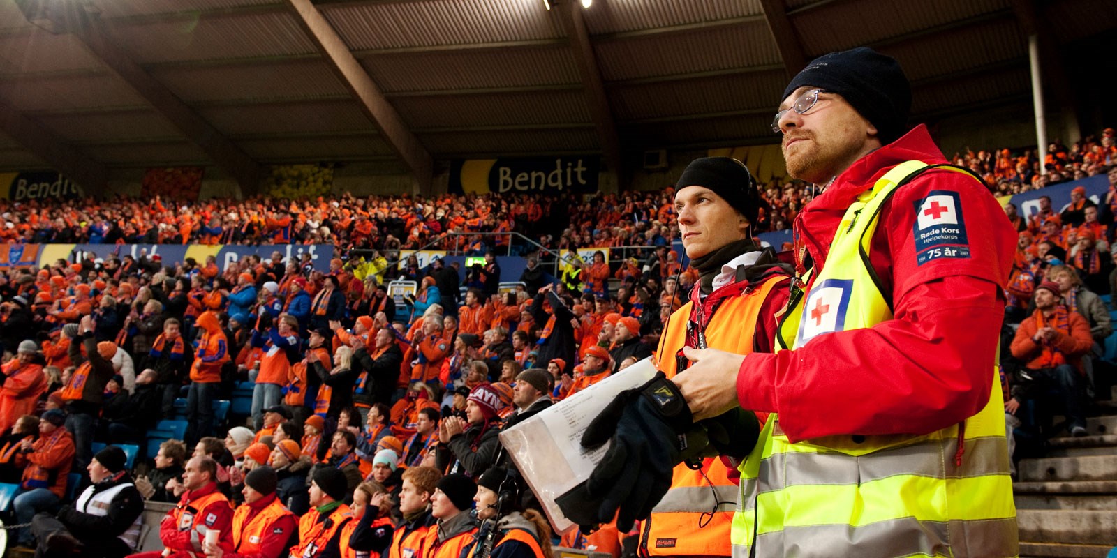 Bilde viser publikum på stadion med 2 personer i front far Røde Kors Hjelpekorps