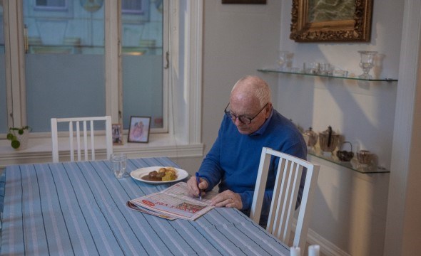 En eldre mann sitter alene ved et spisebord og løser kryssord.