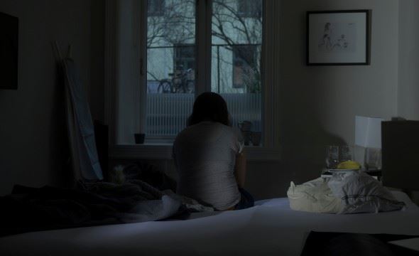 En person sitter på en seng i mørket og ser ut av et vindu med ryggen til kamera.