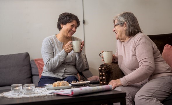 En voksen dame sitter ved siden av en eldre dame i en sofa. De holder begge en kaffekopp mens de smiler og ser på hverandre.