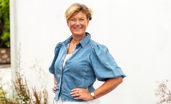 En kvinne ser mot kamera med hendende på hoftene og smiler. Hun har på seg en dongerioverdel.