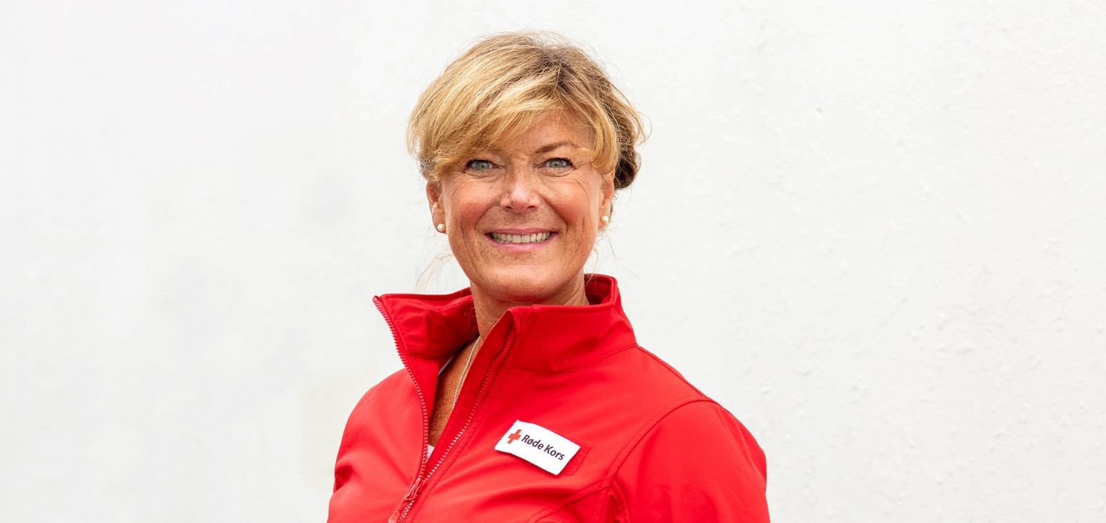 En kvinne med lyst kort hår smiler til kamera. Hun har på seg en rød jakke med logoen til Røde Kors.