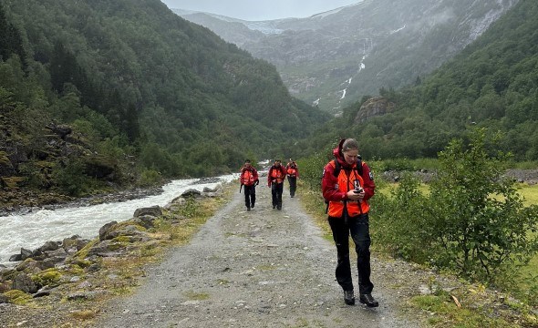 Frivillige går bortover en vei gjennom et dalføre og leter etter en savnet person. Været er grått og skydekket lavt.
