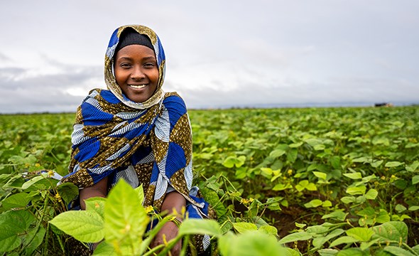 En kvinne med blå hijab sitter ute i en åker og smiler blant de grønne plantene.