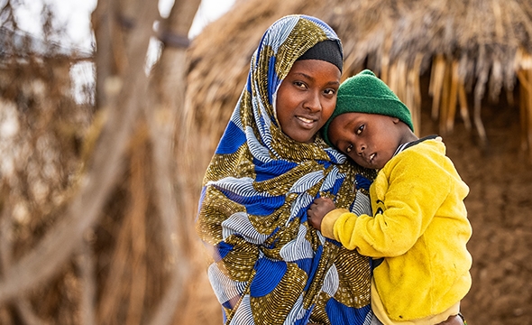 En kvinne med hijab står utendørs med et lite barn i armene sine.