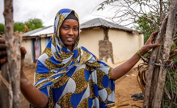 En kvinne med blå hijab står utenfor et hus i en landsby og smiler.