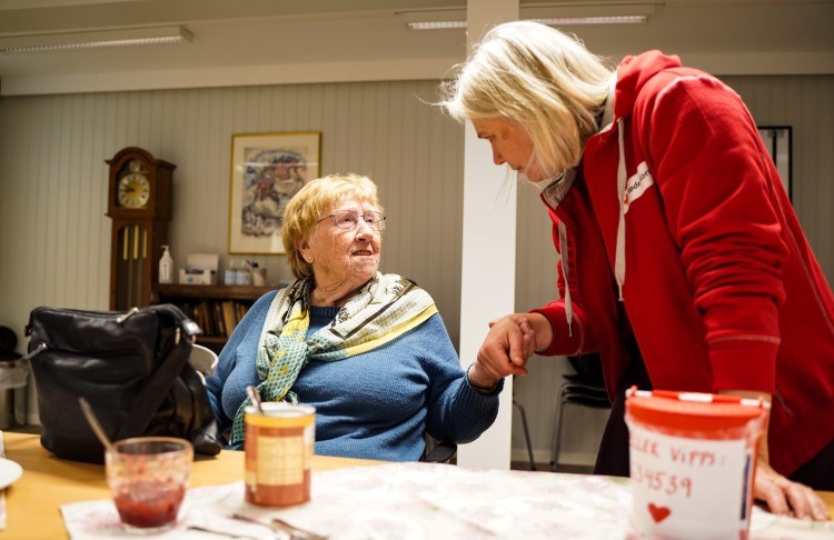 En eldre kvinne sitter på en stol og snakker med en kvinne fra Røde Kors - de holder hverandre i hånden