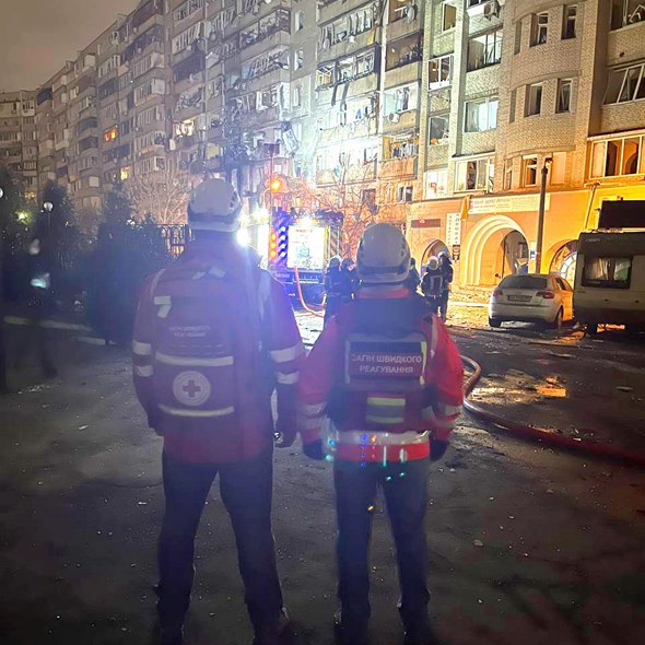 To personer med Røde Kors-uniform og hjelper står foran en bygning som er skadet av krigshandlinger.