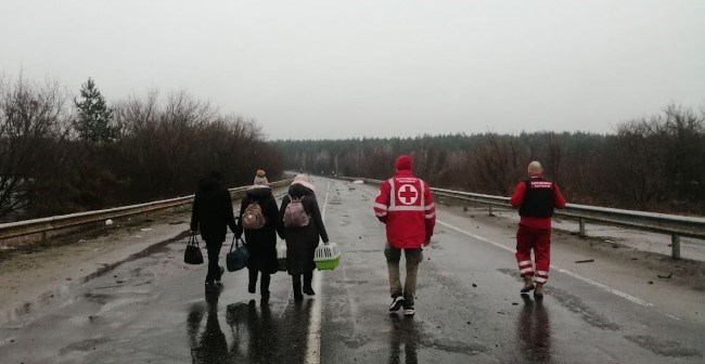 Tre sivile går med to representanter fra Røde Kors langs en vei med bagasje og dyrebur.