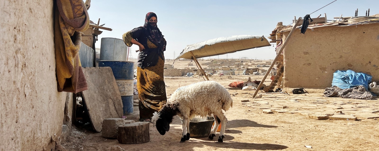 En kvinne står utenfor et enkelt hus sammen familiens eneste sau. Landskapet rundt er varmt og tørt.