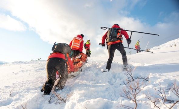 Tre personer fra Hjelpekorps frakter en person på båre opp en bratt snødekket bakke