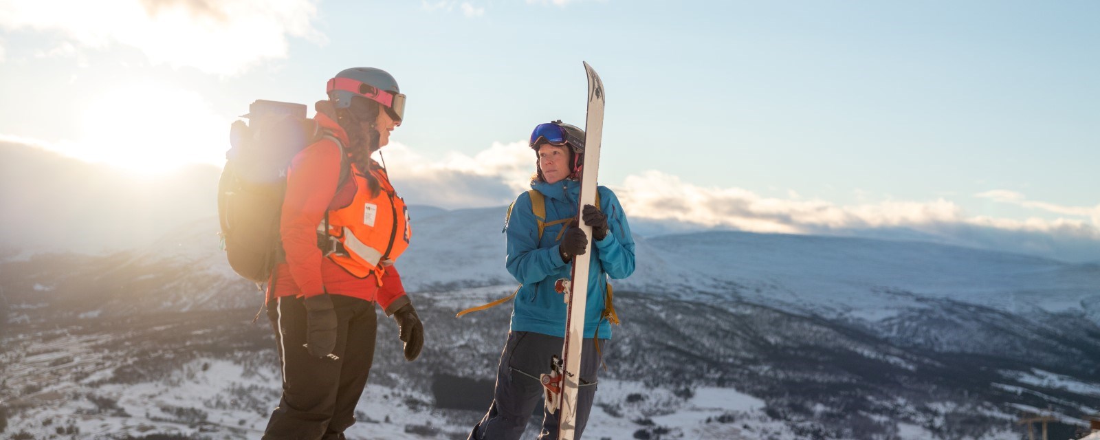 En kvinne fra hjelpekorpset i samtale med kvinnelig turgåer som holder et par fjellski. Snødekt fjell, opphold