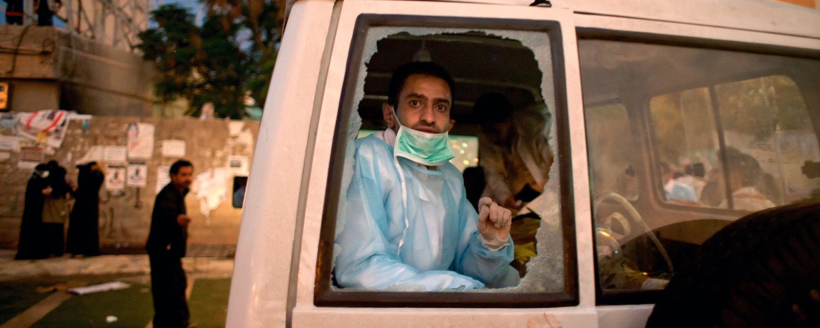En lege sitter i en bil og kikker ut av et knust vindu.