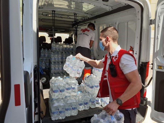 En mann fra Røde Kors bærer ut kasser med vannflasker ut av en bil.