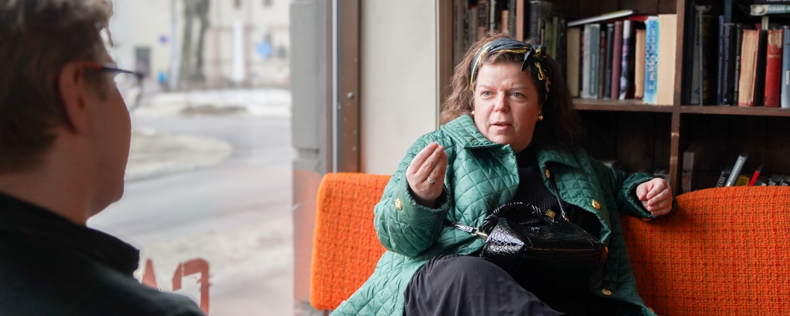 En kvinne med grønn jakke sitter i en oransje sofa på kafé og prater med en mann.
