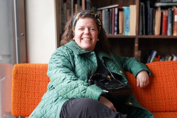 En smilende kvinne med grønn jakke sitter i en oransje sofa på en kafé.