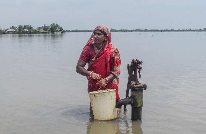 En kvinne står med vann til livet ved siden av en vannpumpe
