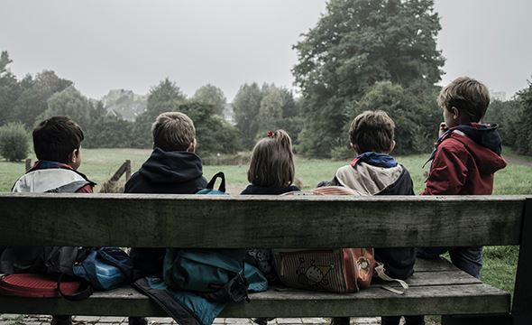 5 barn sitter på en benk med ryggen til. trær og landskap i bakgrunnen