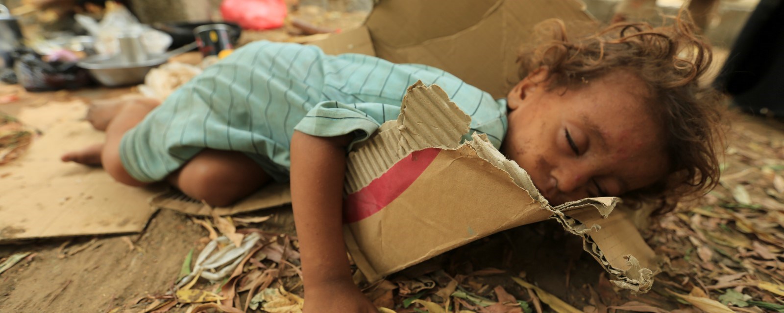 En jente ligger og sover på en pappeske. I bakgrunnen et bål med en kjele