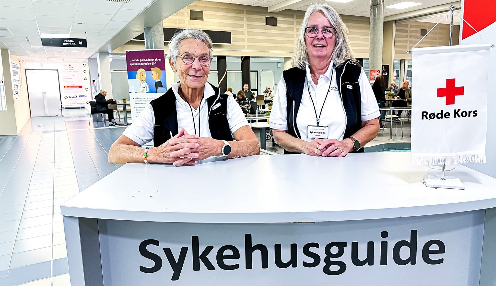 Inne i sykehuset foran Sykehusguide bordet, bak bordet står det to eldre kvinner som er frivillige i Sykehusguide tjenesten.