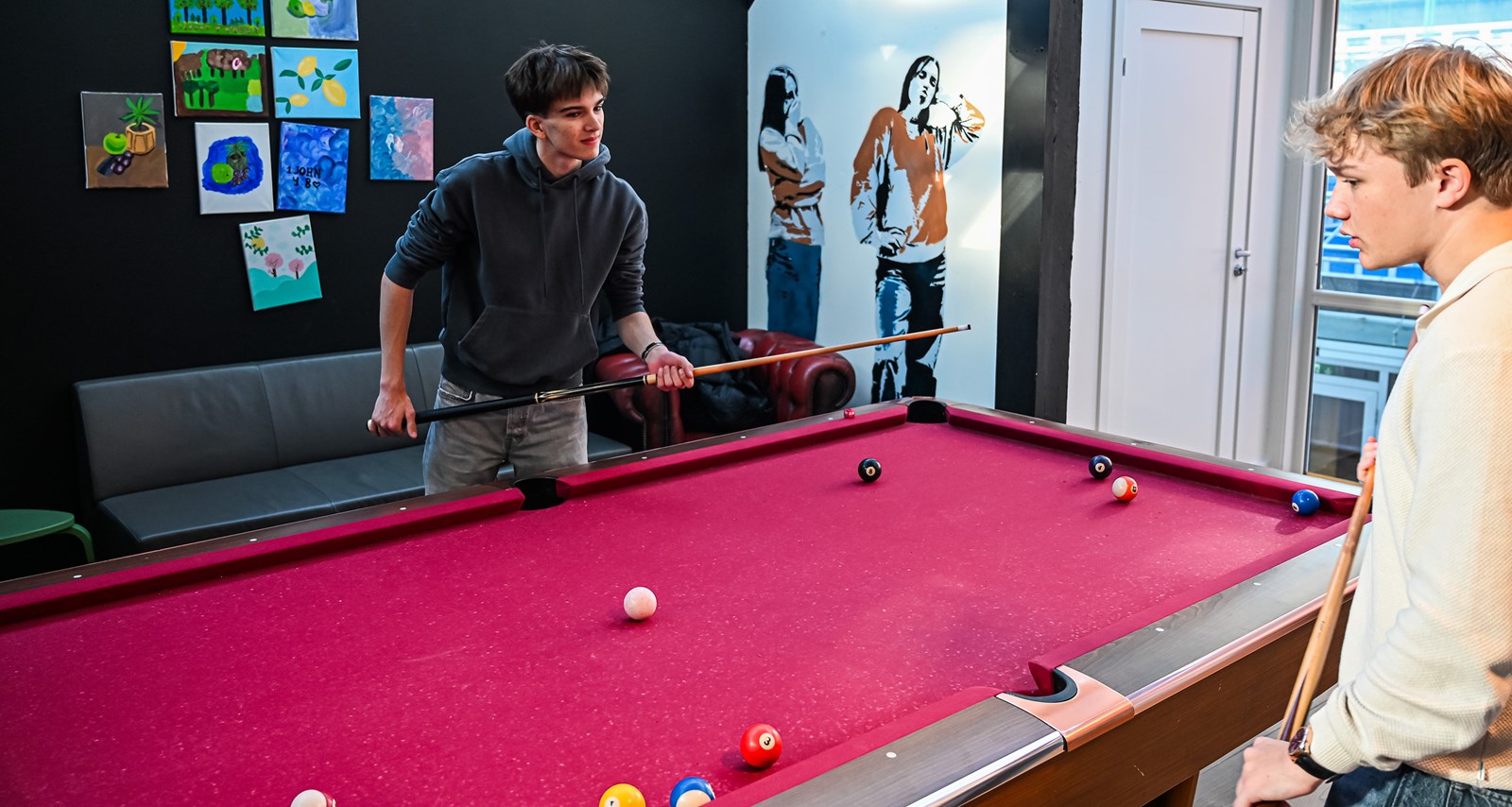 To unge personer spiller biljard på Fellesverket i Kristiansand, et aktivitetshus for ungdom. Kunstverk på veggen og et sosialt miljø understøtter samhold og aktivitet blant deltakerne.
