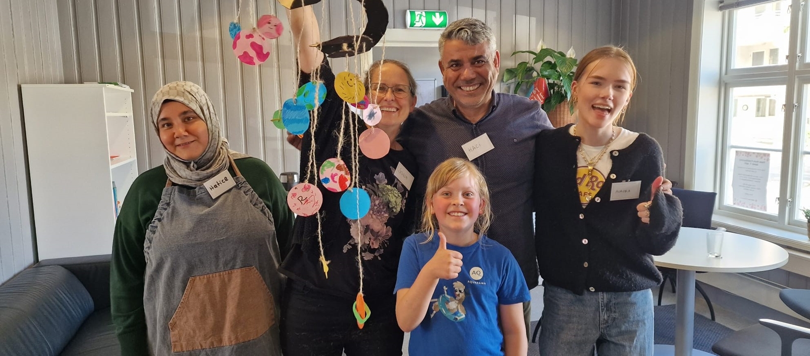 Gruppe mennesker, inkludert barn og voksne, poserer sammen med et fargerikt håndlaget kunstverk laget under en aktivitet på Røde Kors Familiesenter i Kristiansand. De holder et dekorativt objekt med papirfigurer, som representerer samhold, kreativitet og fellesskap.