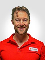 Portrettbilde av en voksen man med kort til middels  lengde med brunt hår, med kort brun blondt skjegg, et stort smil. Har også på seg røde kors uniform.