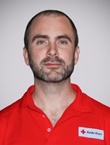 Portrettbilde av en voksen man med utrolig kort mørkt hår, med kort en blanding av mørkt og grått skjegg, et nøyetralt eller passivt smil. Har på seg røde kors uniform.