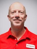 Portrettbilde av en voksen norsk mann uten hår men med grå skjeggstubber, også med røde kors uniformen på.