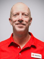 Portrettbilde av en voksen norsk mann uten hår men med grå skjeggstubber, også med røde kors uniformen på.