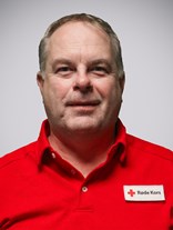 Portrettbilde av en voksen mann med kort brunt hår, hvor delevis av det har blitt grått og er glattbarbert, han har også på seg røde kors uniform.