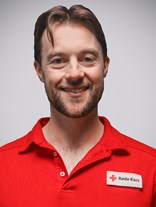 Portrettbilde av en voksen man med kort til middels  lengde med brunt hår, med kort brun blondt skjegg, et stort smil. Har også på seg røde kors uniform.