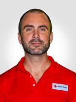 Portrettbilde av en voksen man med utrolig kort mørkt hår, med kort en blanding av mørkt og grått skjegg, et nøyetralt eller passivt smil. Har også på seg røde kors uniform.