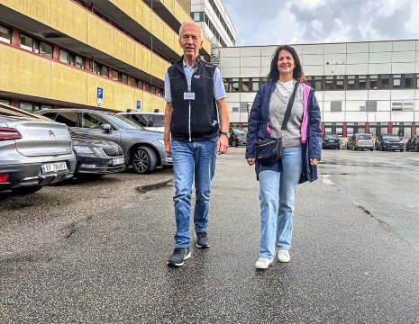 To personer går mot sykehuset på Røde Kors, med parkeringsplasser og moderne bygning i bakgrunnen. Godt vær og avslappet atmosfære.