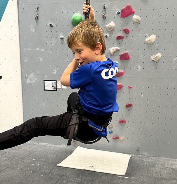 Gutt i klatresele svinger i tau foran en innendørs klatrevegg, under en aktivitet arrangert av Røde Kors sitt familiesenter.