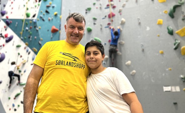 To personer smiler til kamera foran en innendørs klatrevegg, mens barn klatrer i bakgrunnen under et familiearrangement i regi av Røde Kors.