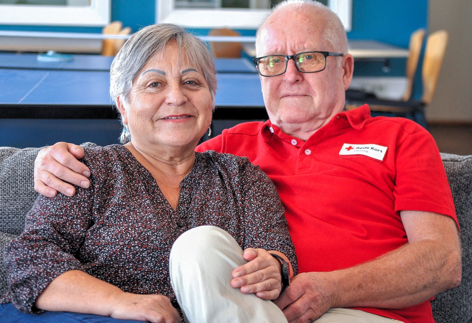 Et eldre par sitter tett sammen på en sofa og smiler mot kamera. Mannen har på seg en rød skjorte med Røde Kors-logo, og de ser avslappede og fornøyde ut.