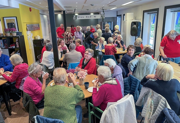 Et rom fullt av eldre mennesker som sitter ved bord og prater sammen under et sosialt arrangement. Flere personer i røde Røde Kors-skjorter hjelper til og serverer kaffe.