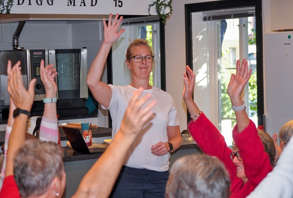 En kvinne i hvit t-skjorte står foran en gruppe eldre mennesker, alle med hendene hevet i været, som del av en felles aktivitet eller avstemning. De befinner seg i et lyst rom med kjøkken i bakgrunnen. Stemningen virker positiv og engasjert.