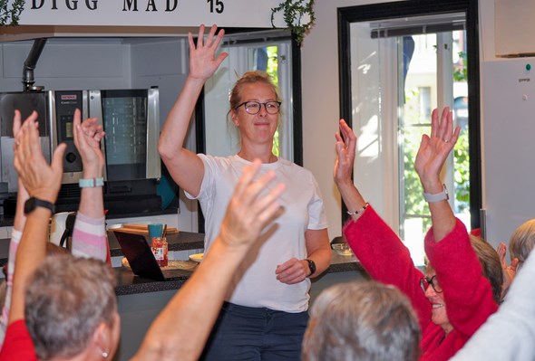 En kvinne i hvit t-skjorte står foran en gruppe eldre mennesker, alle med hendene hevet i været, som del av en felles aktivitet eller avstemning. De befinner seg i et lyst rom med kjøkken i bakgrunnen. Stemningen virker positiv og engasjert.