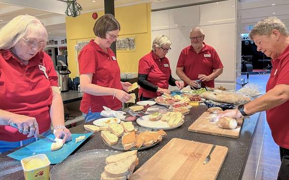 Fem frivillige i røde skjorter forbereder mat sammen på et kjøkken. De smører brød, skjærer ost og legger opp pålegg som ost, skinke og frukt på fat. Stemningen virker hyggelig og samarbeidende.