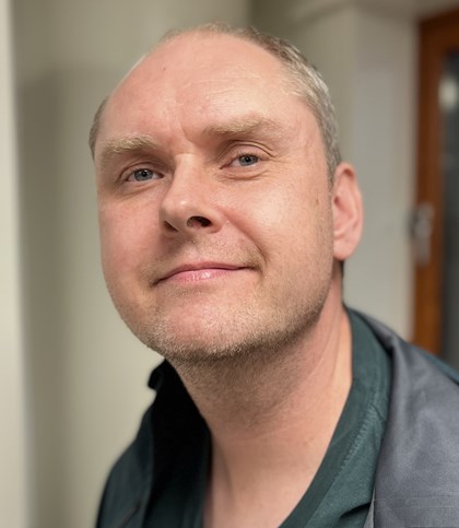 Portrettbilde av en voksen mann med litt lite hår på toppen av hode. Han er ny barbert, har et smil og har på seg en skjorte og jakke.