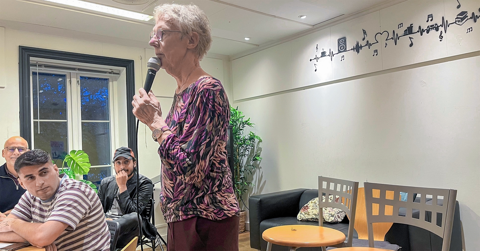 En eldre kvinne står og holder mikrofon mens hun snakker til en gruppe menn som sitter og lytter. Aktiviteten foregår i et møte- og oppholdsrom.